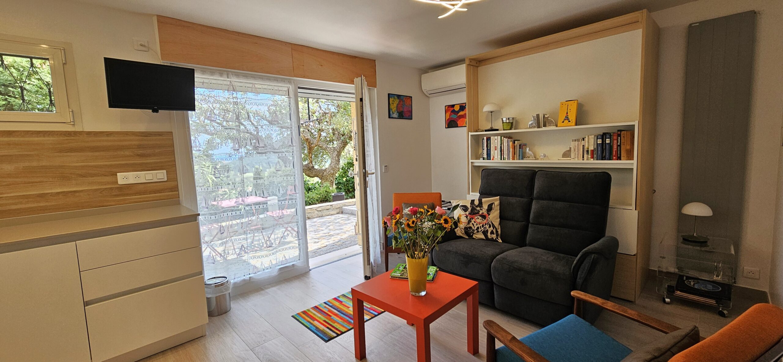 Entrée et salon du rez-de-jardin. Appartement 2 pièces totalement indépendant Entrée et salon du rez-de-jardin. Appartement 2 pièces totalement indépendant. Canapé en tissu gris antracite et bibliothèque ; climatiseur. Porte vitrée donnant accès à l'extérieur et jardin.