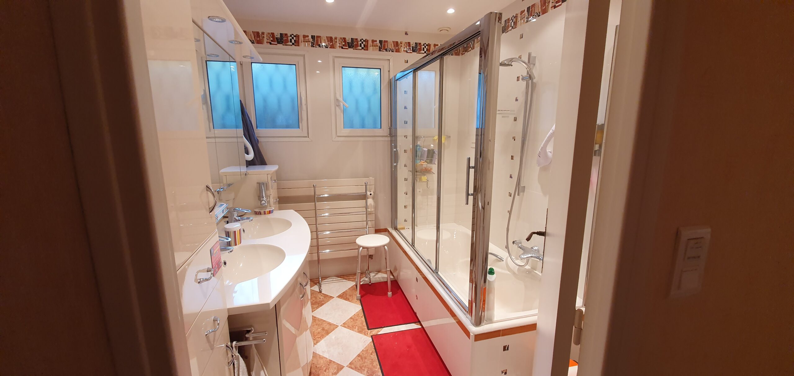 Photo prise de la porte d'entrée de la salle de bains du rez-de-chaussée. Meuble avec double vasque sur la gauche, et sur la droite la baignoire avec un pare-bain coulissant. Au fond, 2 petites fenêtres et en dessous, un radiateur